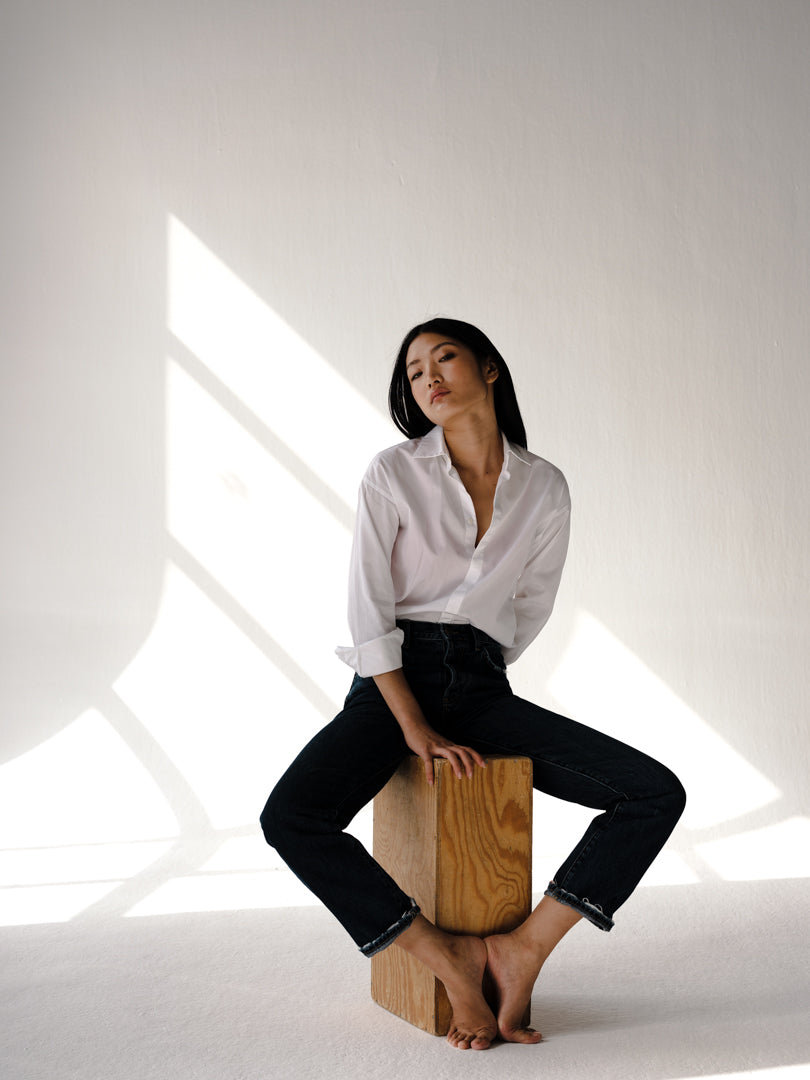 Fit model donning the B O R O デニム (BORO DENIM) 'Eagle Birds' mid blue wash Tokyo jeans, gracefully seated on a wooden cube. The image captures the jeans' blend of relaxed sophistication and modern-day allure.