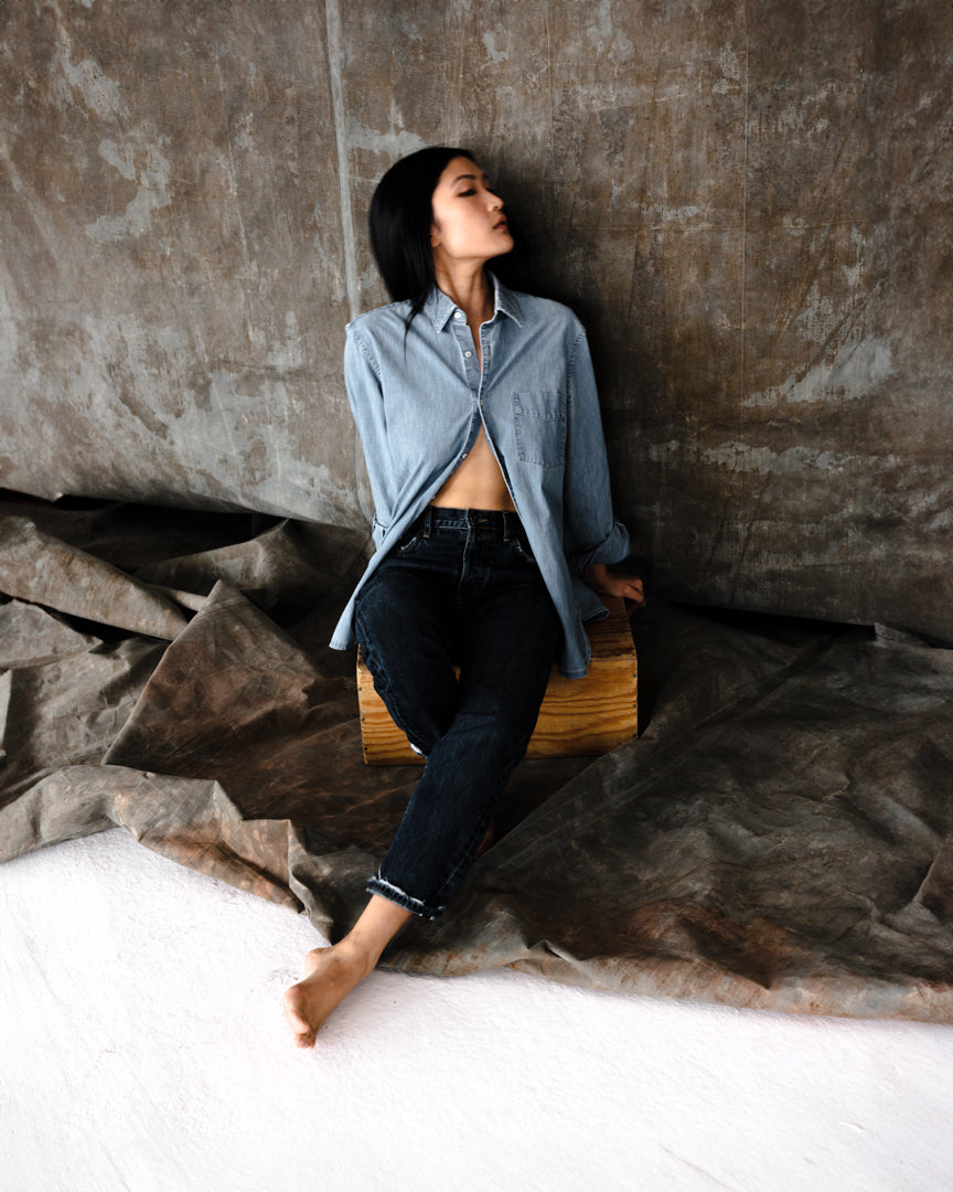 Fit model donning the B O R O デニム (BORO DENIM) 'Eagle Birds' mid blue wash Tokyo jeans, gracefully seated on a wooden cube. The image captures the jeans' blend of relaxed sophistication and modern-day allure.