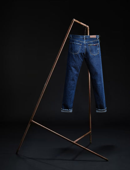Tokyo jeans by B O R O デニム (BORO DENIM) in the 'Eagle Birds' colorway, displayed elegantly on a rack. The back side of the jeans emphasizes the superior Japanese Selvedge fabric and the genuine mid-blue vintage wash, contrasting beautifully with a black backdrop.